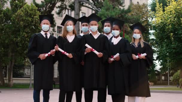Draußen im College-Park posiert eine Gruppe multirassischer Absolventen mit Schutzmaske in der Pandemie des COVID 2019 vor der Kamera und hält Diplome in der Hand. — Stockvideo