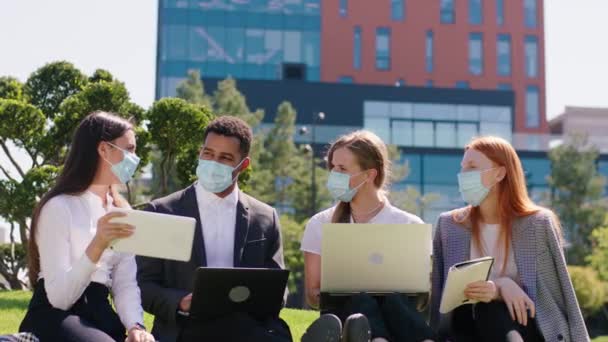 Coronavirus 2019 salgınında çok ırklı bir grup öğrenci kolej derslerinden sonra dizüstü bilgisayar ve tablet kullanarak parkta birlikte çalışıyor koruyucu maskeler takıyorlardı. — Stok video