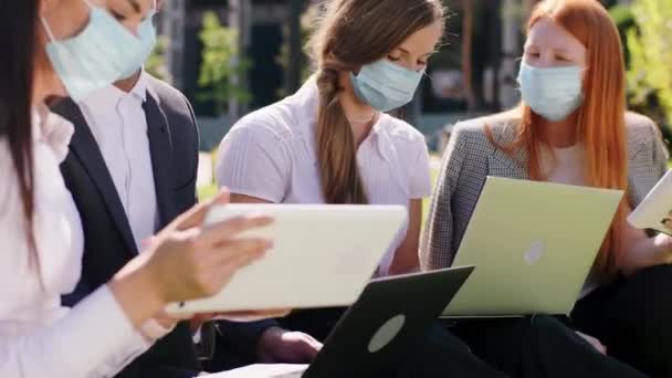 Przed kamerą badają grupę wielorasowych studentów na laptopie i tabletach noszących maskę ochronną w pandemii Koronawirusa w środku parku na trawie. 4k — Wideo stockowe