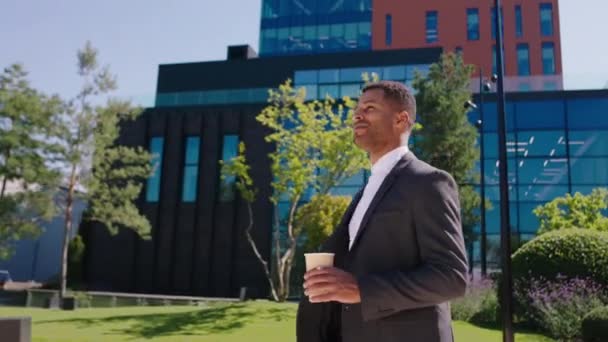 Gestionnaire de bureau attrayant homme noir heureux de marcher pour se rendre au travail à pied à côté des bâtiments modernes du centre d'affaires il tient une tasse avec du café — Video