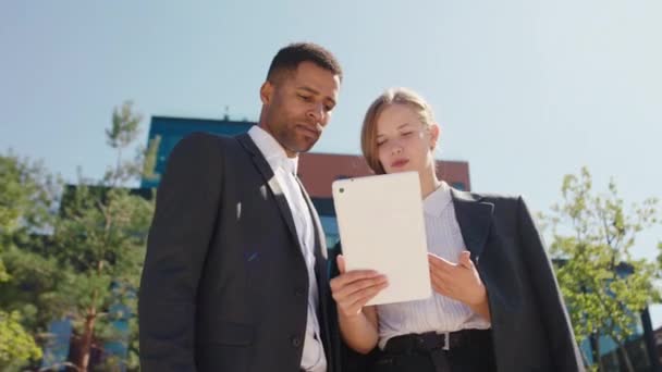 Devant la caméra belle dame gestionnaire de bureau et homme noir employé de bureau discuter ensemble quelque chose en utilisant une tablette pour analyser sur l'arrière-plan architecture de bâtiment moderne — Video