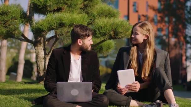Στο σύγχρονο πάρκο κάτω στο γρασίδι όμορφη κυρία και χαρισματικός τύπος δύο μαθητές έχουν ένα έργο μαζί που μελετούν από φορητό υπολογιστή και tablet — Αρχείο Βίντεο