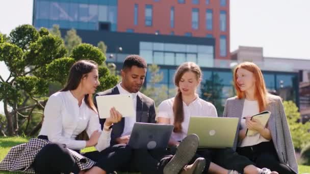 Grup entuziasmat de studenți multirasiale în parc în jos pe iarbă studiind împreună folosind laptopuri și tablete pentru a face un proiect pentru obiect colegiu — Videoclip de stoc