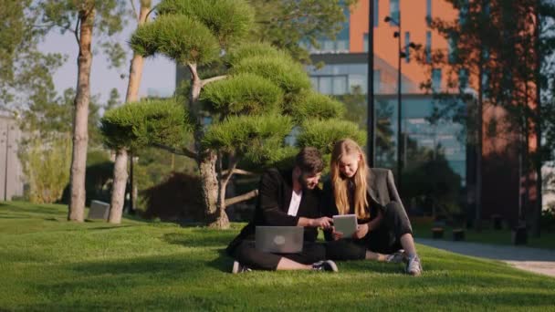 Os estudantes novos olhando bons do par têm um tempo da ruptura fazem exame de um assento no ar fresco para baixo na grama no parque da faculdade que usando laptops e tabuleta a prestar atenção a algo — Vídeo de Stock