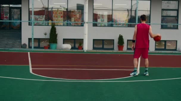 На сучасному стадіоні молодий чоловік грає в баскетбол він кидає м'яч у кошик, він носить червоний спортивний костюм — стокове відео