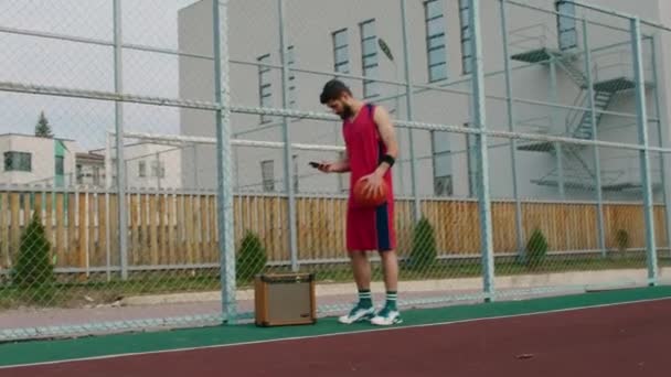 Boisko do koszykówki młody człowiek w czerwonym stroju sportowym przygotować się do gry włożył trochę muzyki na głośnik i zacząć grać z piłką — Wideo stockowe
