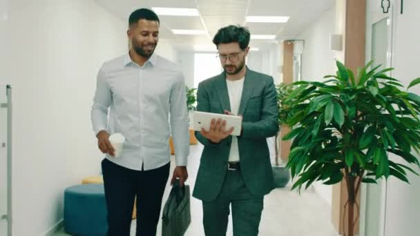 Het is een zeer drukke dag op kantoor, twee werkende mannen in pakken lopen door de gangen en praten terwijl ze een tablet vasthouden, ze passeren veel verschillende mensen — Stockvideo