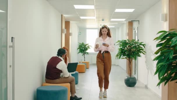 Una hermosa mujer con una camisa rosa y pantalones marrones de moda está caminando por el pasillo de la oficina con su teléfono en la mano y saludando a la gente en el pasillo — Vídeo de stock