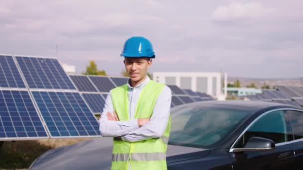 Video primer plano bien parecido hombre asiático ingeniero ecológico posando delante de la cámara al lado de coche eléctrico y grandes paneles solares granja — Vídeo de stock