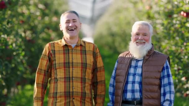 Vor der Kamera posieren fröhlicher und aufgeregter Sohn und Vater Astbauer vor der Kamera und lächeln aufgeregt, um die Ernte vom Obstgarten zu arrangieren — Stockvideo
