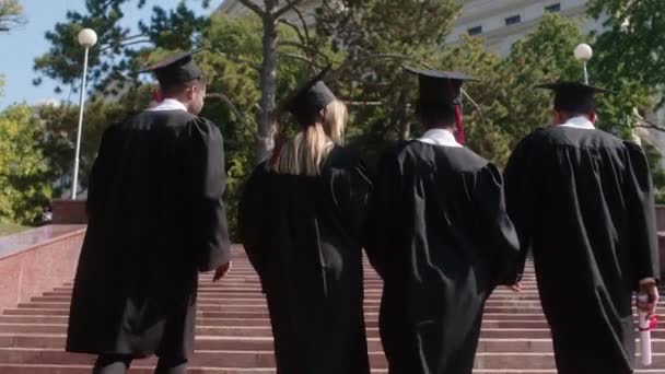 Feliz grupo de estudiantes multirraciales graduados con gorras de graduación subiendo las escaleras fuera del edificio de la universidad después de la graduación. 4k — Vídeos de Stock