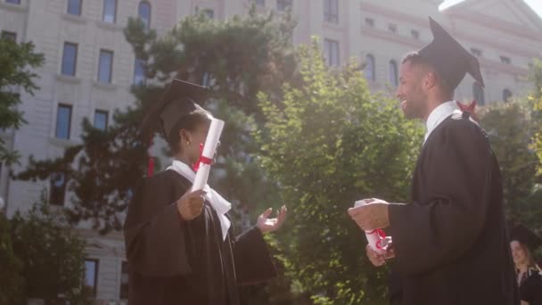 Concept d'obtention du diplôme deux jeunes étudiants charismatiques noir regardant homme et femme discuter après qu'ils aient été diplômés et détenant des diplômes dans le jardin du collège — Video