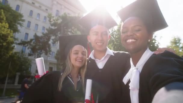 Grupo de estudantes multirraciais graduados duas senhoras e um cara tomando selfie vídeo com a câmera sorrindo grande após a formatura — Vídeo de Stock