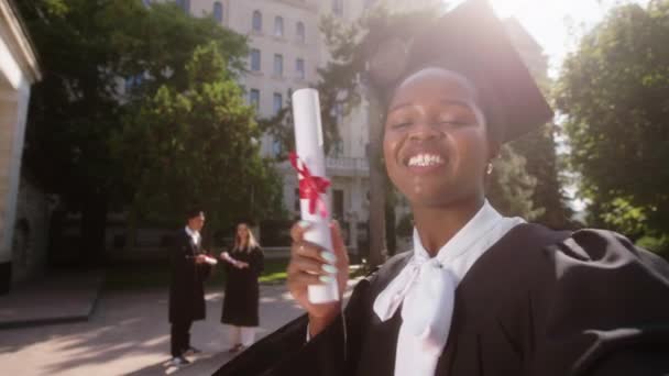 Carismático hermosa mujer negra estudiante graduado hacer un video selfie con cámara después del final de la graduación ella sonriendo gran diploma de celebración en las manos en el jardín de la universidad — Vídeos de Stock