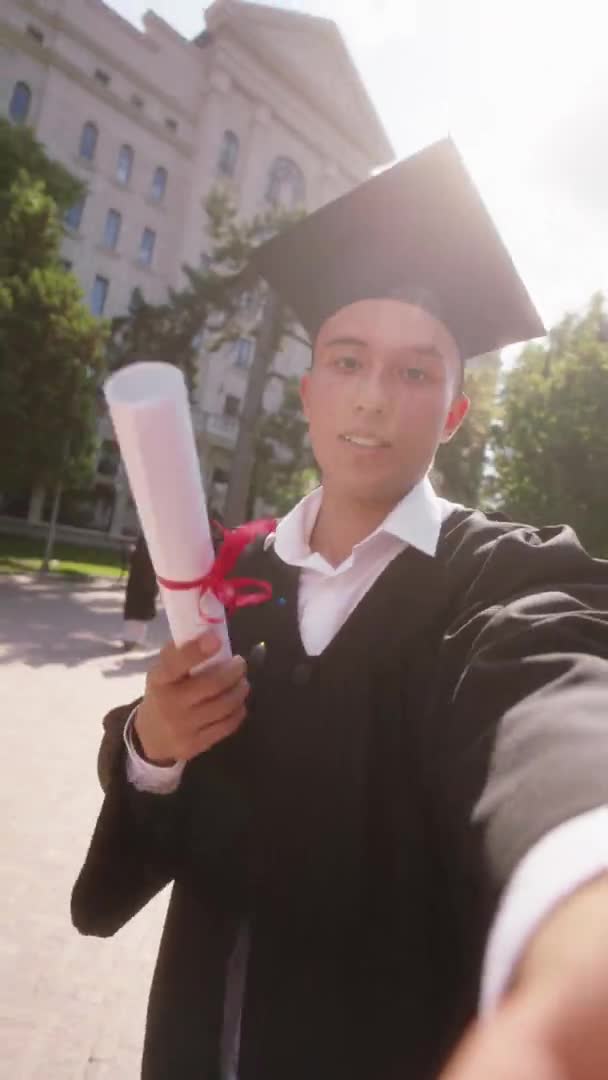 Студент-выпускник с помощью фотоаппарата снимает на видео селфи, очень взволнованный, он выступает перед камерой и показывает диплом, снятый на видео. — стоковое видео