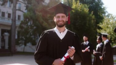 Güneşli bir günde, modern üniversite bahçesinden mezun olmuş bir adama poz verdirerek doğrudan kameraya bakıyor ve elinde diplomasıyla gülümsüyor.