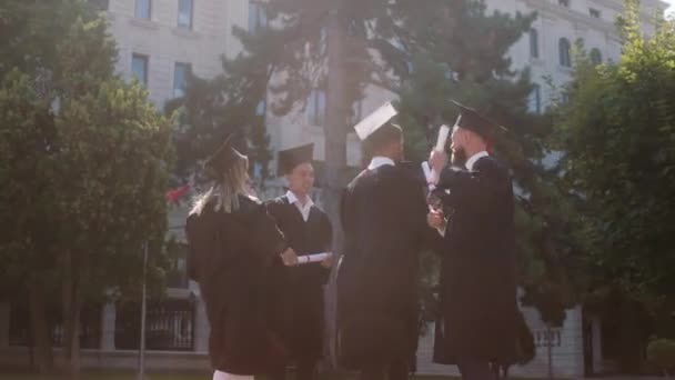 Gran día para un grupo de estudiantes graduados multirraciales con trajes de graduación y gorras que bailan y se sienten emocionados después de tomar los diplomas — Vídeo de stock
