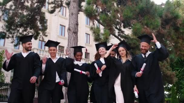 Lachende grote afgestudeerden studenten in de universiteitstuin allemaal samen in de groep voor de camera lopen samen en gooi de afstuderen caps zeer enthousiast — Stockvideo