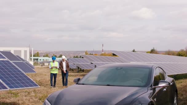 Caminhando na frente da câmera dois engenheiros ecológicos carismáticos discutindo juntos no meio de painéis solares modernos usarem capacetes de segurança — Vídeo de Stock