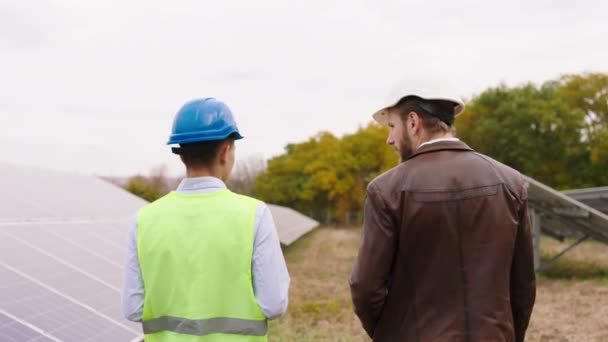 Grünes Energiekonzept auf dem Solarmodulbauernhof mit Video von zwei ökologischen Arbeitern, die durch die Photovoltaik-Batterien gehen und über etwas diskutieren, das sie mit Schutzhelmen tragen — Stockvideo