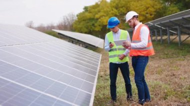 Güneş panellerindeki iki ekoloji mühendisi fotovoltaik pilleri kontrol ediyor ve koruyucu miğfer konsepti olan elektrikli yenilik hakkında bilgi yazmak için dijital tablet kullanıyorlar.