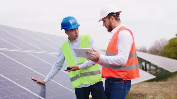 Ingenieros ecológicos multiétnicos en la central de energía que controlan las baterías fotovoltaicas de la tableta digital juntos usan cascos de seguridad — Vídeo de stock