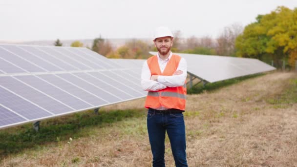 Attractive ecological engineer man at modern solar panels farm posing in front of the camera crossing hands and wearing safety helmet — Stock Video