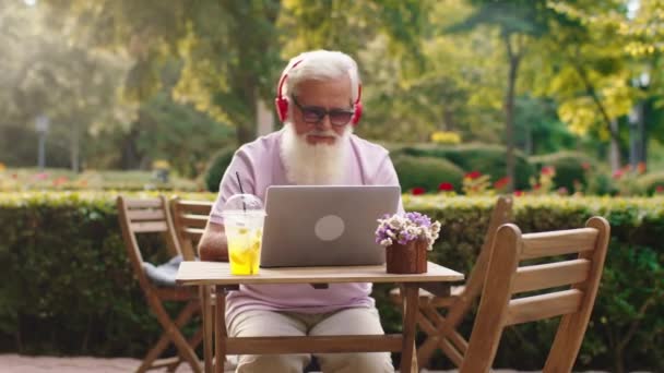 Atraktivní stařec s bílou bradou v kavárně v parku poslouchá hudbu ze sluchátek a pracuje i na notebooku — Stock video