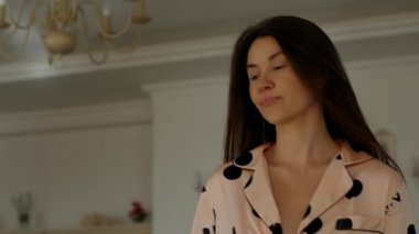Yumuşak pembe benekli pijama giyen genç bir esmer kız bir odada yürürken saçları zarif bir şekilde yüzünün üzerine düşüyor.