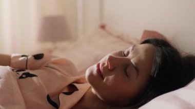 Çok güzel esmer bir kız pembe köpek pijaması giyiyor ve yavaşça yüzünde bir gülümsemeyle uyanıyor. ARRI Alexa Mini 'de çekilen.