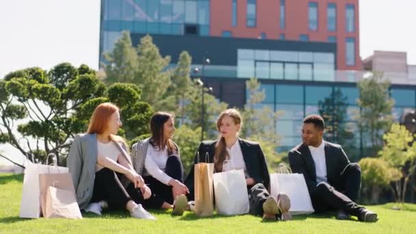 Groupe multiethnique d'amis après un bon shopping prendre un siège sur l'herbe au milieu de la ville bâtiment moderne sur le fond — Video