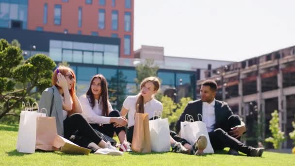 In a modern town in the middle of a large green field sitting down on the grass group of charismatic and multiracial students after a good shopping day they unboxing the shopping bags — Stock Video