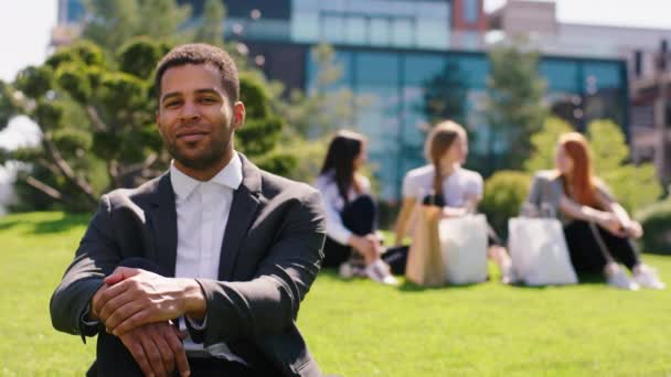 Όμορφος υπάλληλος γραφείου άντρας μαύρος που ποζάρει μπροστά στην κάμερα χαμογελώντας την ώρα του γεύματος στη μέση του μοντέρνου κήπου άλλοι συνάδελφοι στο παρασκήνιο συζητούν μαζί — Αρχείο Βίντεο
