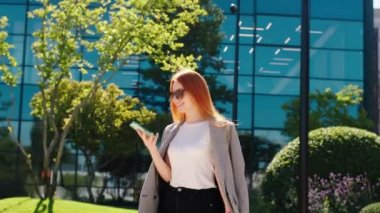 Modern iş binası güneş gözlüklü güzel gülümseyen bayan akıllı telefonunu alıp güneşli bir günde biriyle konuşuyor. 4k