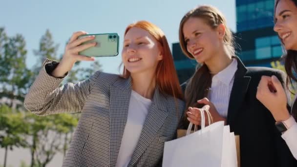 ショッピングバッグを持ちながらスマホの前でポーズをとりながら、笑顔の女性たちが素敵なショッピングセラピーの後に写真を撮りながら、カリスマ性のあるカメラの近くに — ストック動画