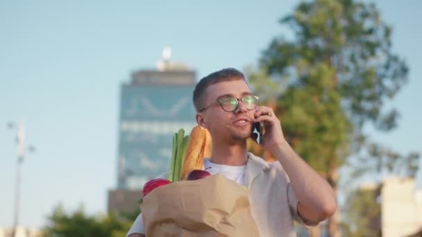 Modern bir sokakta genç bir adam caddede yürüyor ve marketten döndükten sonra elinde sebze ve meyve dolu bir çantayla akıllı telefonla konuşuyor. — Stok video
