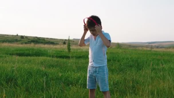 Carismático menino de seis anos pegue seus fones de ouvido e comece a correr pelo campo verde no meio da natureza e aproveite o momento e a infância — Vídeo de Stock