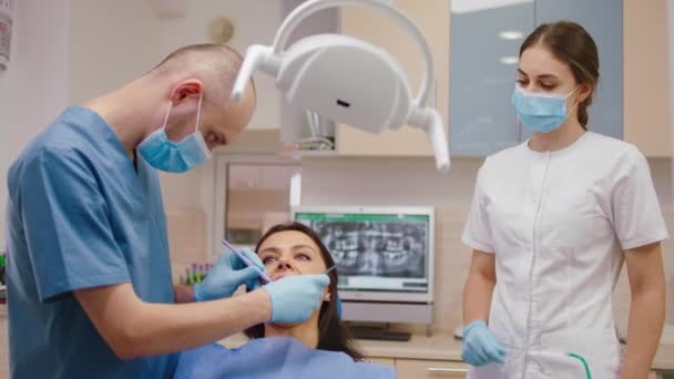 Fiatal fogorvos férfi van egy orális vizsgálat a női beteg egy modern fényes fogászati klinikán a női asszisztens néz óvatos, hogyan fogorvos dolgozik — Stock videók