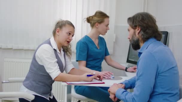 Moderna clínica luminosa paciente maduro tener una conversación con el asistente de la clínica antes de iniciar un chequeo de salud al mismo tiempo médico mujer que trabaja en el ordenador — Vídeo de stock