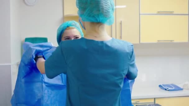Kamera kliniği asistanının önünde cerrahi bir operasyona başlamadan önce doktor için tam korumalı ekipman giymeye yardım ediyor. — Stok video
