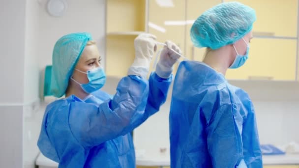 Asistentă medicală de sex feminin în clinică înainte de a începe o operație chirurgicală ajutând la îmbrăcarea medicului pe echipamentul special. 4k — Videoclip de stoc