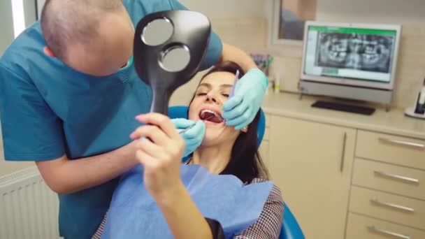 Красивая терпеливая женщина с идеальной улыбкой и белыми зубами у стоматолога возьмите зеркало, чтобы посмотреть результаты лечения — стоковое видео