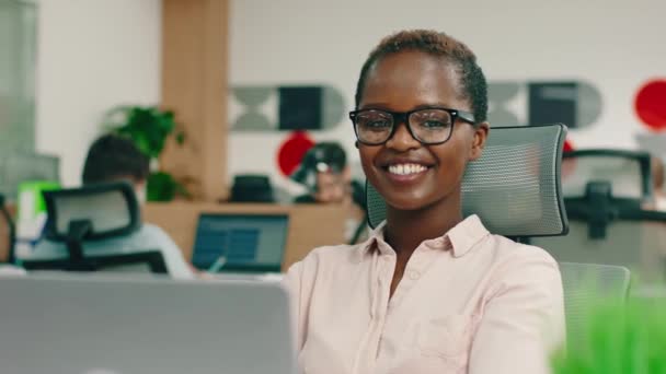 Eine sehr klug aussehende schwarze Frau mit kurzen Haaren trägt eine Brille und eine Bluse, sie sitzt in einem Büro und sieht sehr glücklich aus, zu arbeiten — Stockvideo