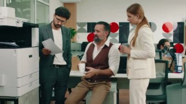 Yaşlı, yakışıklı bir iş adamı, iki iş arkadaşıyla konuşup onlara danışmanlık yaparken kızıl saçlı bir kadın, modern görünümlü bir ofiste asistanlarıyla yaptıkları iş hakkında konuşuyor.