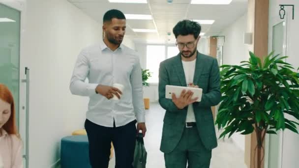 Zwei sehr gut aussehende und hingebungsvoll aussehende Manager spazieren durch das Büro und reden über die Arbeit, einer hält ein Tablet in der Hand, während der andere eine Aktentasche in der Hand hält, sie grüßen die Leute — Stockvideo