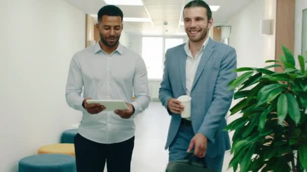 Двоє розумних відомих бізнесменів йдуть через офіс і розмовляють, вітаючи всіх, вони сидять за круглим столом з синім диваном, щоб поговорити про роботу — стокове відео