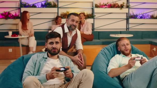 턱수염을 가진 세 남자가 콩 백 의자에 앉아 비디오 게임을 하며 신나게 서로를 응원하고 있습니다 — 비디오