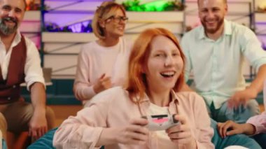 Kızıl saçlı bir kız video oyunları oynuyor ve etrafındaki bir grup insan neşeyle gülüyor.