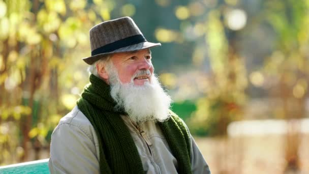 Knappe oude man met een vintage hoed dicht bij de camera poserend buiten in het park gaat hij op stoel zitten — Stockvideo