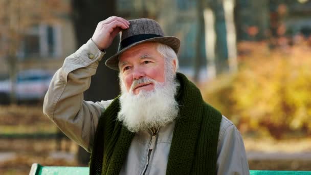 Attraktiver alter Mann mit Vintage-Hut im Park nimmt den Hut ab und legt ihn dann zurück, während er im Spätherbst sitzt — Stockvideo
